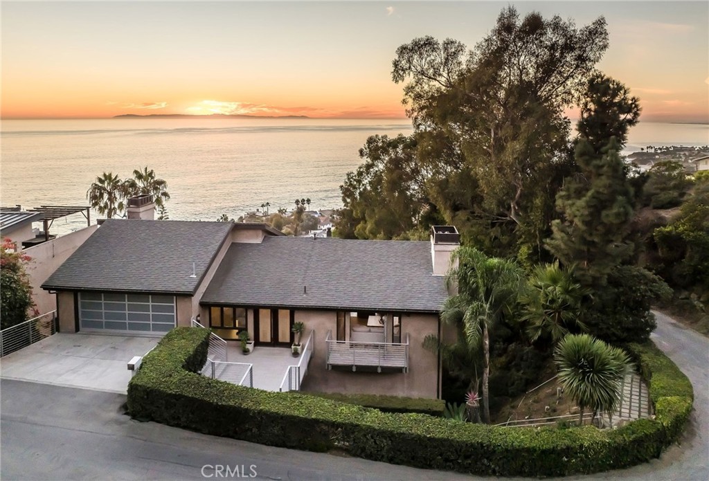 A contemporary, private coastal estate nestled into a hillside with awe-inspiring panoramic views of the Pacific Ocean, Catalina Island, and the city lights. The interiors of this home offer a pinnacle of current design, with high-end finishes and voluminous living spaces. Located off the entryway, the formal living room, anchored by a sleek fireplace, is flanked by accordion glass doors, allowing in bountiful light and ocean breezes. The great room comprises a more casual sitting area and an updated kitchen, offering the coveted open floor plan. Great for gatherings as well as relaxation, this space features soaring ceilings, a second fireplace, and access to a large deck with expansive views. Clean lines extend into the fully modernized kitchen with a central island, ample storage, a premium suite of stainless steel appliances, and access to the dining area for seamless entertaining. The adjacent balcony is large enough to accommodate a seating area, serving as an extension of the indoor entertaining space. The sleeping quarters are located on the lower level. An ocean-facing primary retreat encompasses a spacious bedroom with a fireplace, a balcony, a spa-like fully equipped bathroom, and a massive walk-in closet. All of the secondary bedrooms are ensuite and could serve as guest accommodations, a home gym, or an office. This property is surrounded by lush, mature landscaping, and its additional features include whole-house Sonos surround sound, Lutron lighting, a security system, an attached two-car garage, and a private driveway. While private, the location is convenient, with easy access to the city’s iconic beaches, the Montage Laguna Beach resort, local restaurants, and shopping.