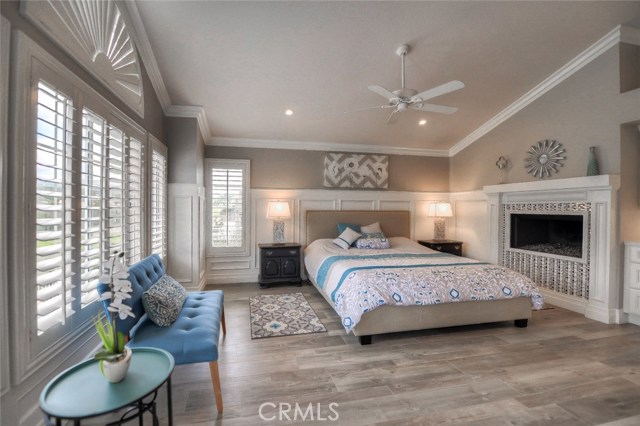 Master bedroom with volume ceiling, gas fireplace, raised panel wainscoting, wood-like tile flooring, crown molding, plantation shutters, and ceiling fan.