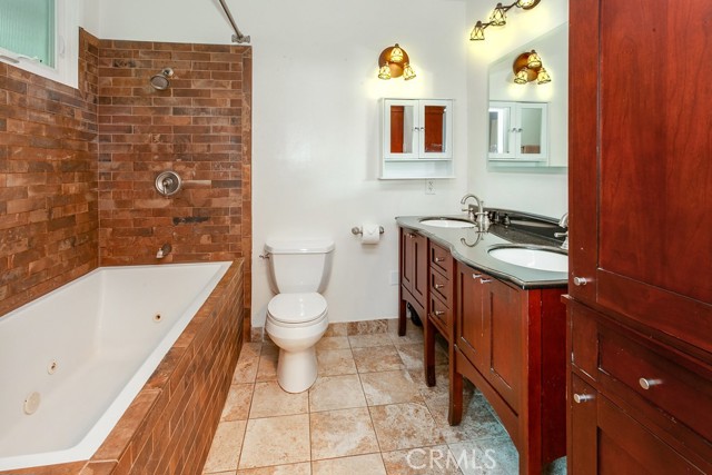Generously sized remodeled private en suite primary bathroom with pocket door, rich wood-grained vanity, dual basins, lighted touchscreen mirror, designer lighting, custom tiled jetted soaking tub/shower and easy-care custom tiled floors.