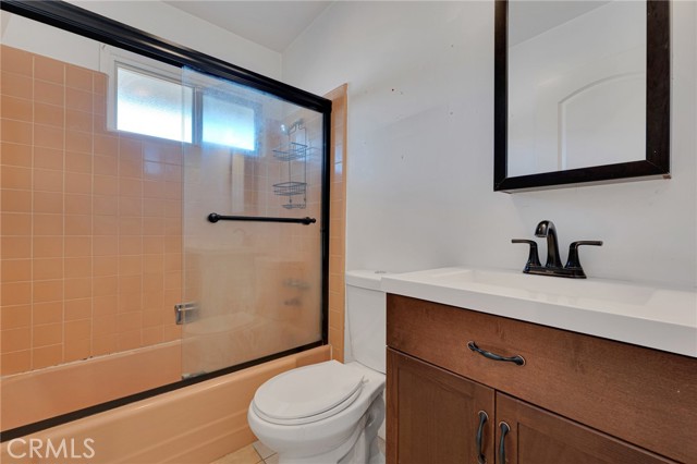 Full bathroom with modern vanity, integrated basin, framed dressing mirror with sundry storage, glass enclosed tiled tub/shower, privacy window and tiled floor.