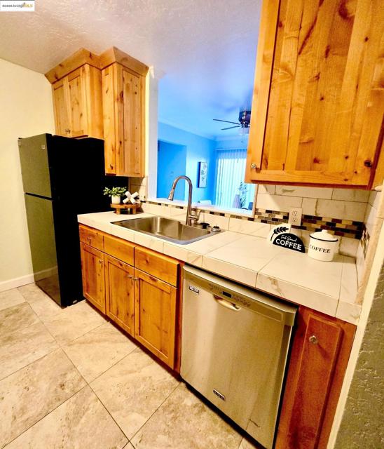 Kitchen with tile counters, freestanding refrigerator, stainless steel dishwasher, decorative backsplash, and a textured ceiling