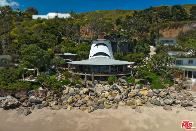 An architectural treasure and an icon of Malibu design with 122 feet of beachfront, Sandcastle was the hand-built home of visionary architect (and surfer) Harry Gesner. Offered for sale for the first time, this tranquil Western Malibu oceanfront property is, according to Gesner, "a dream place, built with love." When the young architect set out to build his family home, he had big ideas, little money, and an abiding passion for the natural beauty of the secluded cove with a nearby surf break and an empty plot right next door to his most famous building, Wave House. With a roofline inspired by a sandcastle, Gesner was determined to build a home that would fulfill what turned out to be a lifelong philosophy: "You have to have a view. You want perspective to your life." The home's texture was determined in part by the materials he salvaged as an early evangelist of sustainable construction: telephone poles, reclaimed bricks, wall panels made from aqueduct pipes, birdseye maple from a high school gym, old-growth redwood harvested in the 1800s, and windows and doors saved from one of Hollywood's silent film theaters. As Gesner said about Sandcastle in Houses of the Sundown Sea: The Architectural Vision of Harry Gesner, the book about his life and work, "The spirit in the design and materials are what you immediately feel in this house. All people who enter and spend a brief period or long time here immediately feel at home and relaxed. The experience never becomes commonplace or boring because over everything is the sound and rhythm of the waves breaking on the shore in front of the house." At the home's entry is a lovely library/sitting area with floor-to-ceiling bookshelves and ocean views. With massive spoke-like beams and a wood-plank ceiling, the heart of the main house is a window-wrapped space that comprises generously scaled living and dining areas with an enormous brick fireplace at its hub. Inspired by the Hollywood Bowl, Gesner built the fireplace as a stage for his wife, actress Nan Martin, with a huge polished concrete hearth. A lovely solarium with stained glass panels flanks the dining area, and the ocean views enjoyed from the interior areas extend to the wraparound deck just outside.Echoing the cylindrical shape of the house itself, the kitchen wraps around a circular island and has tile countertops, stained glass lunettes fitted into the beams overhead, a fireplace, updated appliances, a breakfast bar, and a walk-in pantry. On the home's main level are two en-suite bedrooms, one with built-ins suitable for an office.Upstairs is the primary suite tower, with high ceilings, huge beams, charming eyebrow windows, a brick fireplace, an ocean-view sitting area, and a spiral staircase with handmade driftwood treads leading up to a studio/loft/meditation space.The property's additional structures include an ocean-view "tree house" apartment with a kitchenette, living room/dining area, bedroom, bath, and wraparound deck; a "boat house" with a full kitchen, ocean-view living/dining room with a built-in table and porthole windows, a full bath, and a bedroom; and another one-bedroom apartment "nest" with ocean view and stained glass above an indoor/outdoor cabana. There is also an open-air living area, deck, and teepee-covered daybed.The approximately 0.73-acre property has a three-car garage and additional parking platform, a covered deck, a brick patio, lawn, an outdoor shower, surfboard and other storage areas, steps to the beach, and multiple vantage points for breathtaking sunset views. Sandcastle, which will be sold as is, has abundant outdoor space. A legacy estate with privacy, incomparable character, and singular style.