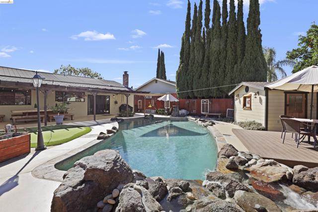 Swimming pool with rock waterfall feature, surrounding concrete patio, wooden deck, and adjacent storage structure