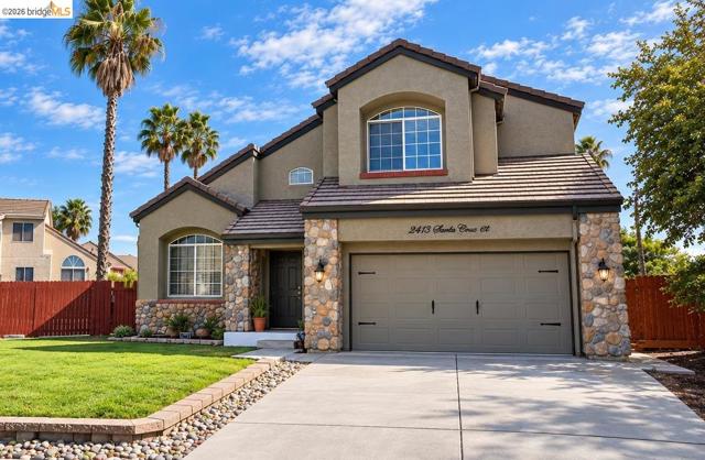 Multi-level stucco exterior featuring stone veneer accents, a tiled roof, concrete driveway, and a manicured lawn with a river rock border