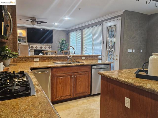 Kitchen with stainless steel appliances, brown cabinets, light stone countertops, crown molding, and a ceiling fan