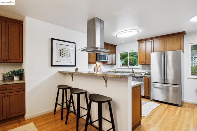 Kitchen featuring appliances with stainless steel finishes, a breakfast bar area, brown cabinetry, light wood finished floors, and island exhaust hood