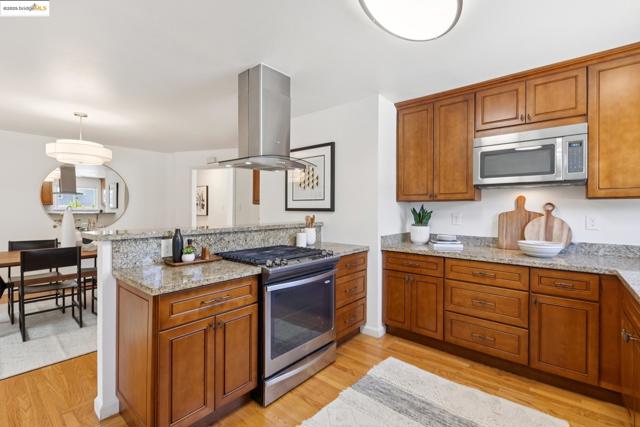 Kitchen with brown cabinetry, stainless steel appliances, light stone countertops, and island exhaust hood