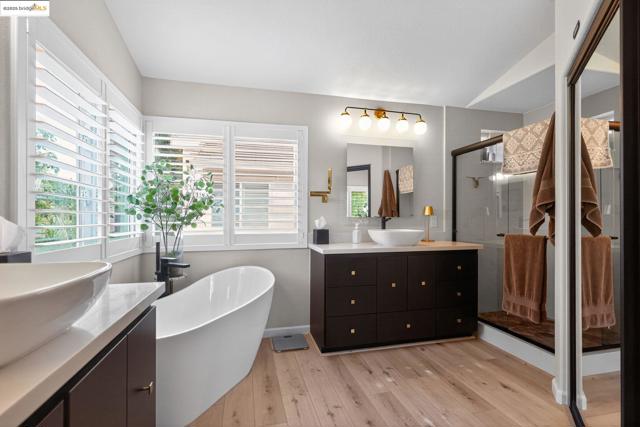 Full bath featuring two vanities, a stall shower, a freestanding tub, light wood-style floors, and vaulted ceiling
