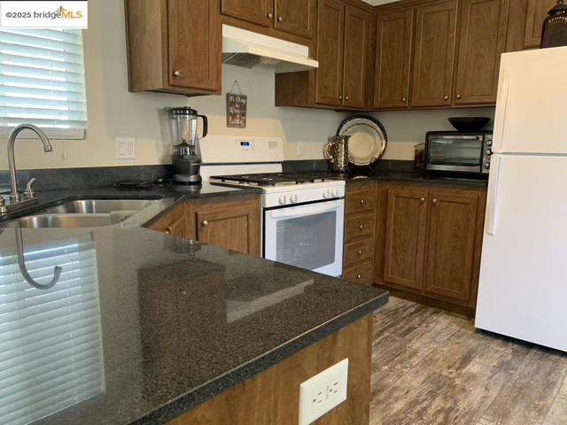 Kitchen featuring white appliances, under cabinet range hood, wood finished floors, and dark stone countertops