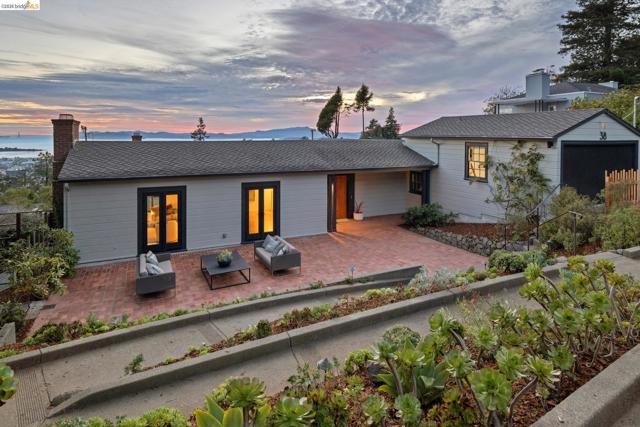 Expansive brick patio, light-colored siding, dark-trimmed double doors, attached garage, and mature landscaping