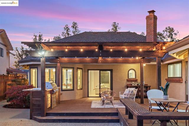 Back of house at dusk featuring an outdoor kitchen, outdoor dining space, a chimney, a wooden deck, and stucco siding
