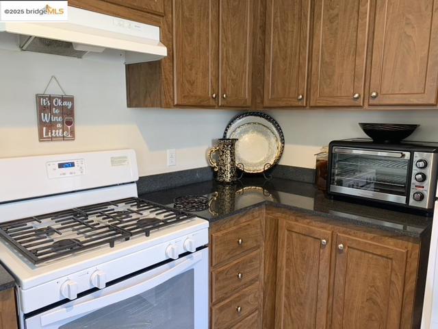 Kitchen with white range with gas cooktop, under cabinet range hood, and dark stone countertops