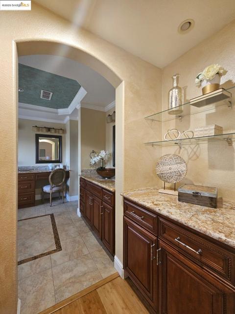 Bar area with open shelves, light stone counters, built in desk, ornamental molding, and a textured wall
