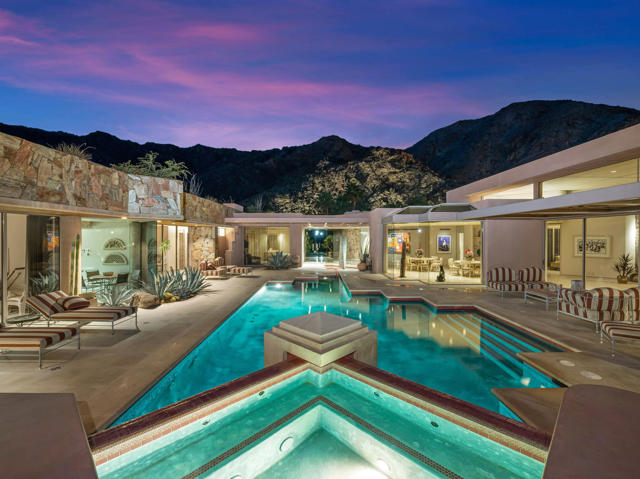 This contemporary styled 7,258 +/- square foot home, designed by acclaimed architects Frank Urrutia and  David Prest, represents a collaboration of creativity. The architects carefully forged this 51,836 +/- square foot privacy lot's natural terrain to craft a habitable dwelling with views centered on the pool, spa and waterways against a mountain backdrop. The landscape design artfully includes a forked waterfall feature tumbling down the mountainside providing a dramatic visual effect along with its water-conserving desert landscape pallet. The sprawling floor plan featuring floor-to-ceiling windows and doors, offers a spacious primary suite wing complimented by four ensuite guest rooms (one currently used as an office). The home offers a solar power grid that helps minimize the costs of maintaining this home. Two of the guest suites are divided by a common sitting room with kitchenette and are accessed separately through the main entry and off the pool area. The design layout offers a large sit-down bar with view window backdrop, a step-down living room with fireplace and a formal dining area. Off the living areas and along the western elevation of the home are relaxing patios and a second spa with vistas of our amazing desert sunsets. The gourmet-style kitchen encompasses a spacious and functional kitchen with center-island, breakfast area and family room with fireplace. The garage is designed to accommodate 3 full size cars plus golf cart.
