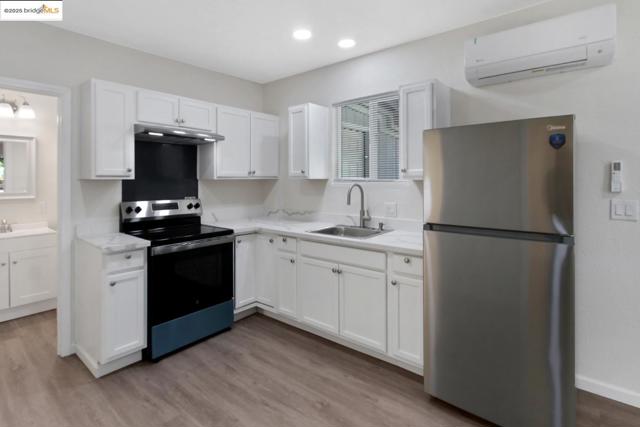 Kitchen with stainless steel appliances, a wall mounted air conditioner, white cabinets, light wood finished floors, and under cabinet range hood